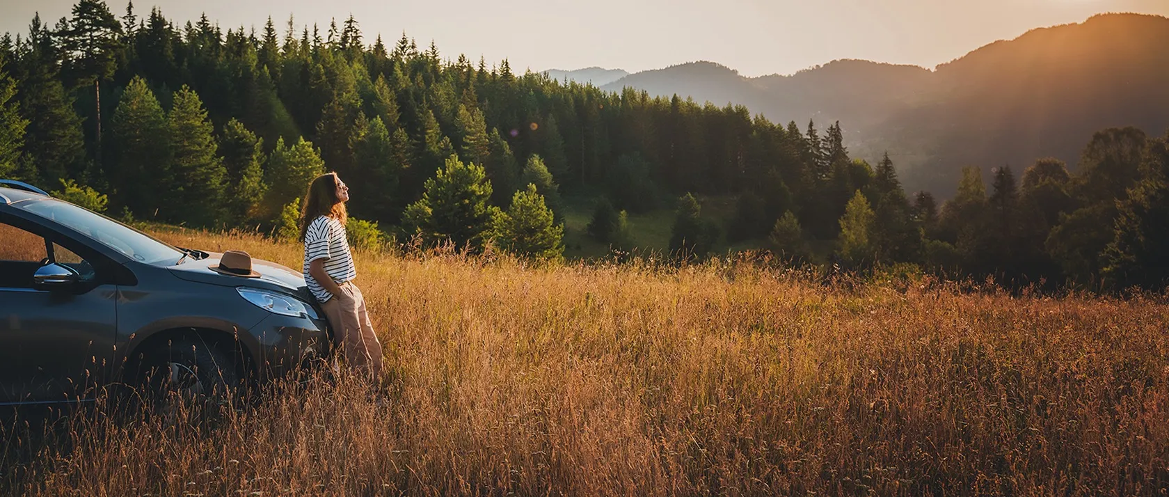 A driver resting against her car in a field at sunset, with forested hills behind